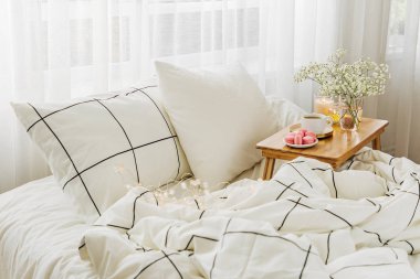Yatakta çiçeklerle birlikte kahve ve mumlardan oluşan ahşap tepsi. Çizgili battaniye ve yastık ile beyaz yatak çarşafları. Yatakta kahvaltı. Hygge kavramı.