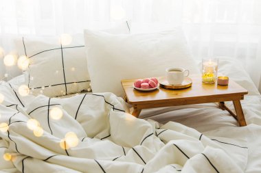 Yatakta ki tahta kahve tepsisi ve mumlar. Çizgili battaniye ve yastık ile beyaz yatak çarşafları. Yatakta kahvaltı. Hygge kavramı.