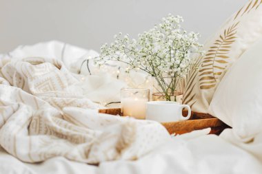 Yatakta çiçeklerle birlikte kahve ve mumlardan oluşan ahşap tepsi. Çizgili battaniye ve yastık ile beyaz yatak çarşafları. Yatakta kahvaltı. Hygge kavramı.