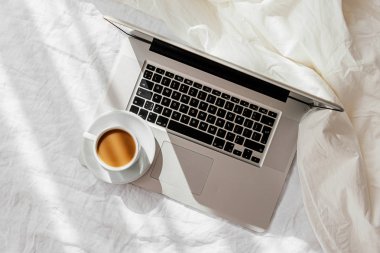 Beyaz yatakta battaniyeli dizüstü bilgisayar ve bir fincan kahve. Evde çalışma konsepti. Sabah ışığı 
