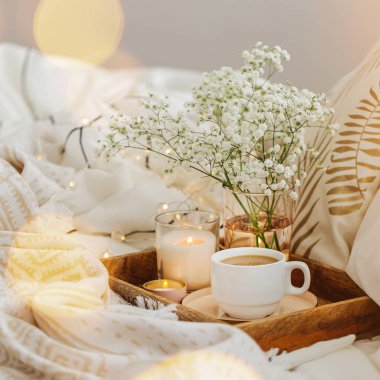 Yatakta çiçeklerle birlikte kahve ve mumlardan oluşan ahşap tepsi. Çizgili battaniye ve yastık ile beyaz yatak çarşafları. Yatakta kahvaltı. Hygge kavramı.