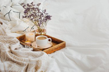 Yatakta çiçeklerle birlikte kahve ve mumlardan oluşan ahşap tepsi. Çizgili battaniye ve yastık ile beyaz yatak çarşafları. Yatakta kahvaltı. Hygge kavramı.