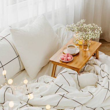 Yatakta çiçeklerle birlikte kahve ve mumlardan oluşan ahşap tepsi. Çizgili battaniye ve yastık ile beyaz yatak çarşafları. Yatakta kahvaltı. Hygge kavramı.