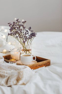 Yatakta çiçeklerle birlikte kahve ve mumlardan oluşan ahşap tepsi. Çizgili battaniye ve yastık ile beyaz yatak çarşafları. Yatakta kahvaltı. Hygge kavramı.