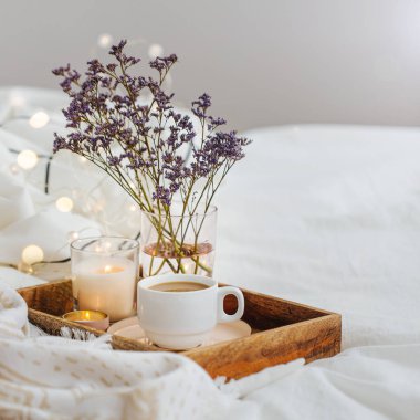 Yatakta çiçeklerle birlikte kahve ve mumlardan oluşan ahşap tepsi. Çizgili battaniye ve yastık ile beyaz yatak çarşafları. Yatakta kahvaltı. Hygge kavramı.