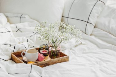 Yatakta çiçeklerle birlikte kahve ve mumlardan oluşan ahşap tepsi. Çizgili battaniye ve yastık ile beyaz yatak çarşafları. Yatakta kahvaltı. Hygge kavramı.
