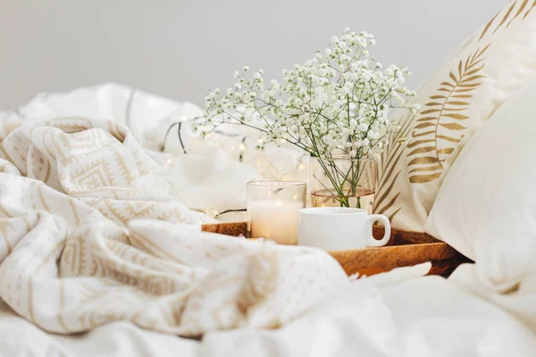 Wooden tray of coffee and candles with flowers on bed. White bedding sheets with striped blanket and pillow. Breakfast in bed. Hygge concept.