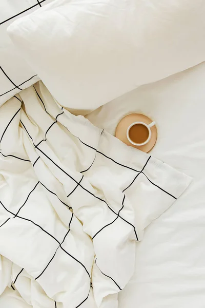 Cup of coffee on bed. White bedding sheets with striped blanket and pillow.