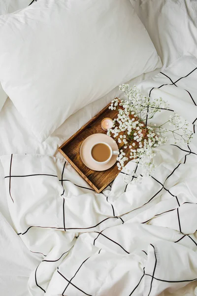 Wooden tray of coffee and candles with flowers on bed. White bedding sheets with striped blanket and pillow. Breakfast in bed. Hygge concept.