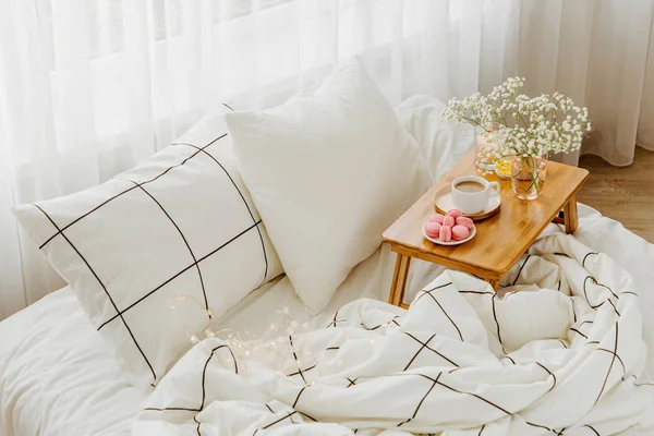 Yatakta çiçeklerle birlikte kahve ve mumlardan oluşan ahşap tepsi. Çizgili battaniye ve yastık ile beyaz yatak çarşafları. Yatakta kahvaltı. Hygge kavramı.