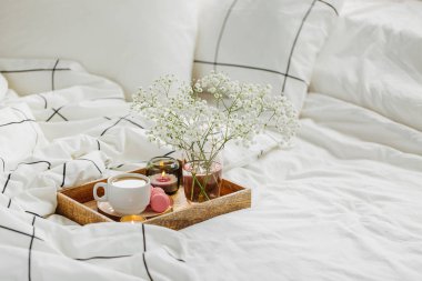 Yatakta çiçeklerle birlikte kahve ve mumlardan oluşan ahşap tepsi. Çizgili battaniye ve yastık ile beyaz yatak çarşafları. Yatakta kahvaltı. Hygge kavramı.