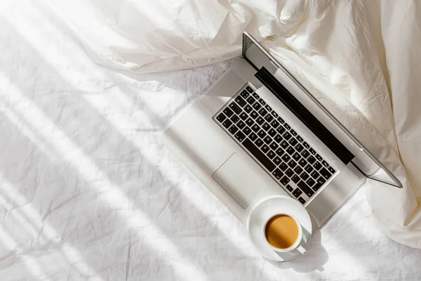 Laptop and cup of coffee on white bed with a  blanket. Work at home concept. Morning light