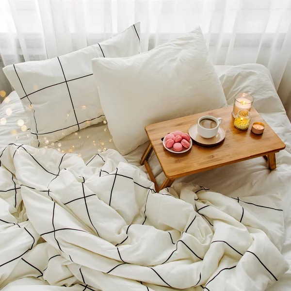 Wooden tray of coffee and candles on bed. White bedding sheets with striped blanket and pillow. Breakfast in bed. Hygge concept.