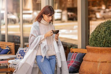 Güzel kız istirahat ve terasta kafede oturan kahve içme, yünlü ekose battaniye sarılı. Sonbahar sabahı.