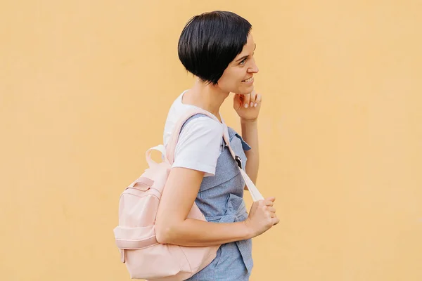 Portrait of female student with a backpack on pale yellow wall.