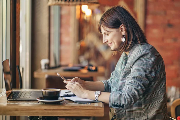 Young business woman in gray blazer using smartphone and working on net-book, sitting in the cafe near the window with coffee. Cozy autumn or winter morning.