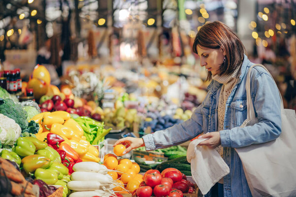 Woman is chooses fruits and vegetables at food market. Reusable eco bag for shopping. Sustainable lifestyle. Eco friendly concept. 