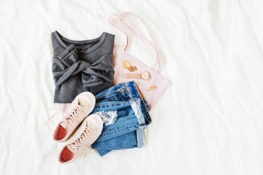 Gri kazak, kot pantolon, eko çanta ve beyaz çarşaf üzerinde yatakta spor ayakkabı. Kadın şık sonbahar veya kış kıyafeti. Modaya uygun giysi kolajı. Düz döşeme, üst görünüm. 