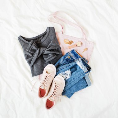 Gri kazak, kot pantolon, eko çanta ve beyaz çarşaf üzerinde yatakta spor ayakkabı. Kadın şık sonbahar veya kış kıyafeti. Modaya uygun giysi kolajı. Düz döşeme, üst görünüm. 