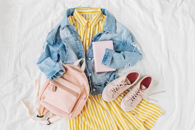 Mavi jean ceket ve sırt çantası, kitap ve beyaz spor ayakkabıları ile Sarı elbise. Kadınların şık sonbahar veya bahar kıyafeti. Üniversite için modaya uygun kıyafetler. Okul konseptine geri dönelim. Düz döşeme, üst görünüm.