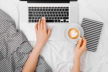 Genç bir kadın evinde, yatağında kahve içiyor ve laptopuyla çalışıyor.