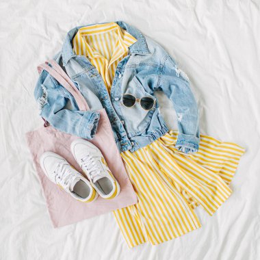 Mavi kot ceket ve beyaz yatakta çanta ve spor ayakkabı ile sarı elbise. Kadınların şık sonbahar veya bahar kıyafeti. Modaya uygun kıyafetler. Moda konsepti. Düz döşeme, üst görünüm.