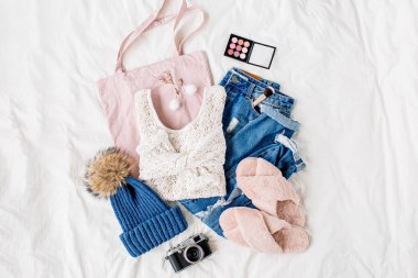 Beyaz örme kazak, mavi kot pantolon, eko çanta ve beyaz çarşaf üzerinde yatakta şapka. Kadın şık sonbahar veya kış kıyafeti. Modaya uygun giysi kolajı. Düz döşeme, üst görünüm. 