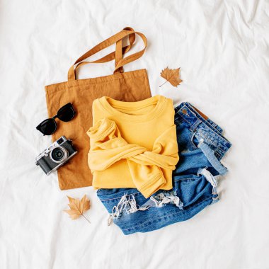 Sarı kazak, mavi kot pantolon, eko çanta ve beyaz çarşaf üzerinde yatakta fotoğraf makinesi. Kadın şık sonbahar veya kış kıyafeti. Modaya uygun giysi kolajı. Düz döşeme, üst görünüm. 