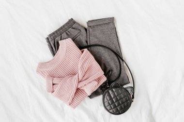 Beyaz levha üzerinde çanta ile soluk pembe sıcak kazak ve gri pantolon. Kadın şık sonbahar veya kış kıyafeti. Modaya uygun giysi kolajı. Düz döşeme, üst görünüm. 