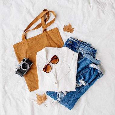 Beyaz gömlek, kot pantolon, eko çanta ve beyaz çarşaf üzerinde yatakta fotoğraf makinesi. Kadınların şık sonbahar kıyafeti. Modaya uygun giysi kolajı. Düz döşeme, üst görünüm. 