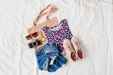 Parlak gömlek, mavi kot pantolon, spor ayakkabı, eko çanta ve beyaz çarşaf üzerinde yatakta fotoğraf makinesi. Kadınların şık sonbahar kıyafeti. Modaya uygun giysi kolajı. Düz döşeme, üst görünüm. 