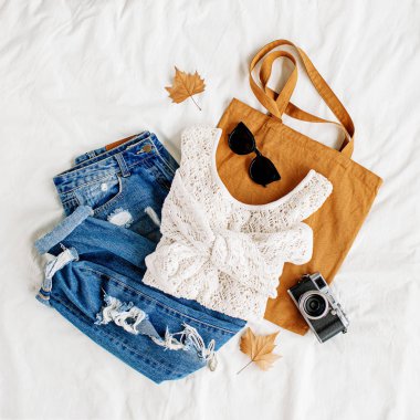 Beyaz örme kazak, kot pantolon, eko çanta ve beyaz çarşaf üzerinde yatakta fotoğraf makinesi. Kadın şık sonbahar veya kış kıyafeti. Modaya uygun giysi kolajı. Düz döşeme, üst görünüm. 