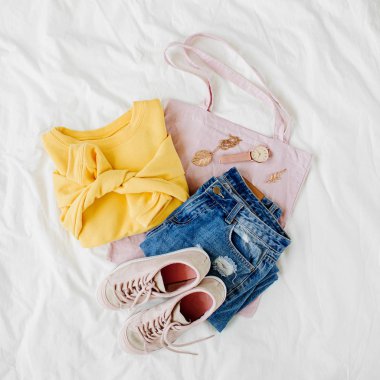 Sarı kazak, mavi kot pantolon, eko çanta ve beyaz çarşaf üzerinde yatakta spor ayakkabı. Kadın şık sonbahar veya kış kıyafeti. Modaya uygun giysi kolajı. Düz döşeme, üst görünüm. 