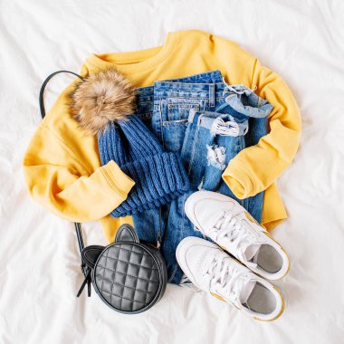 Rahat sarı kazak, kot pantolon, spor ayakkabı ve beyaz çarşaf üzerinde yatakta şapka. Kadın şık sonbahar veya kış kıyafeti. Modaya uygun giysi kolajı. Düz döşeme, üst görünüm. 