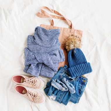 Rahat mavi kazak, kot pantolon, spor ayakkabı ve beyaz çarşaf üzerinde yatakta şapka. Kadın şık sonbahar veya kış kıyafeti. Modaya uygun giysi kolajı. Düz döşeme, üst görünüm. 