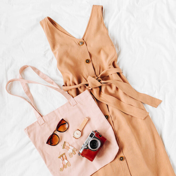 Beige dress with eco bag and accessories on white bed. Women's stylish autumn or summer outfit. Trendy clothes. Flat lay, top view.