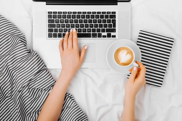 Genç bir kadın evinde, yatağında kahve içiyor ve laptopuyla çalışıyor.
