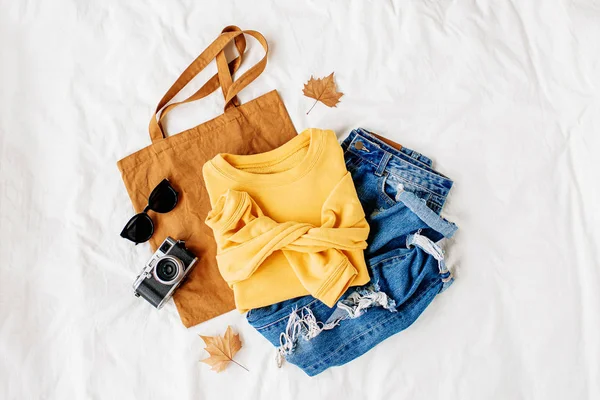 黄色毛衣 蓝色牛仔裤 环保袋和相机在床上的白色床单 女士时尚的秋冬套装 时髦的服装拼贴画 顶视图 — 图库照片