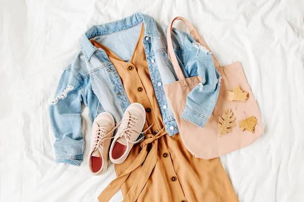 蓝色牛仔裤夹克和米色连衣裙 白色床上带手提包的运动鞋 女装时尚的秋装 时髦的衣服 顶视图 — 图库照片