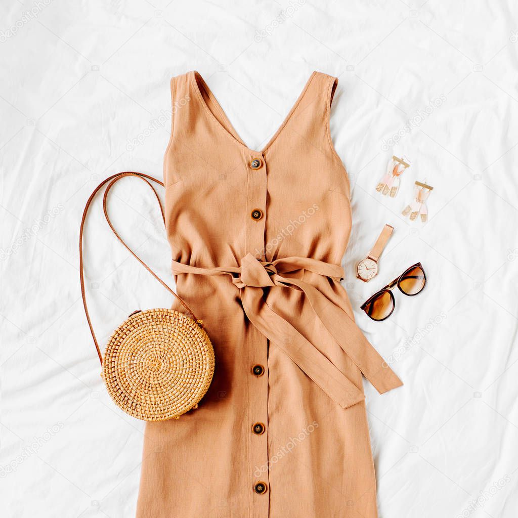 Beige dress with bamboo bag and accessories on white bed. Women's stylish autumn or summer outfit. Trendy clothes. Flat lay, top view.