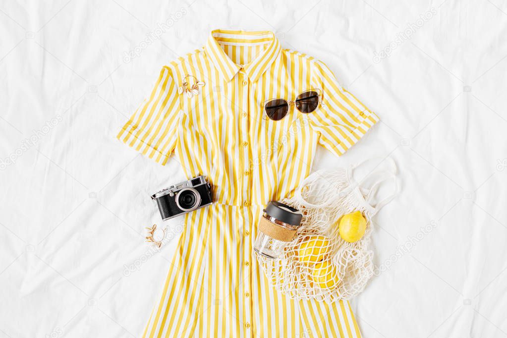 Yellow dress with stripes with eco bag, sunglasses and photo camera on white bed. Women's stylish  summer outfit. Trendy clothes. Vacation concept.  Flat lay, top view.