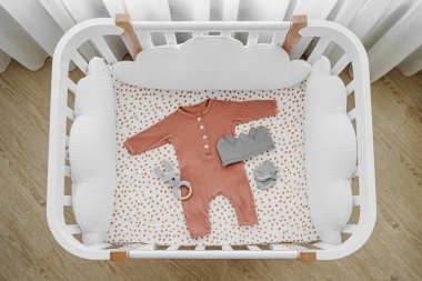 Bebek odasında yastık şeklinde bulutlar olan beyaz ahşap bebek beşiği. Yeni doğan kıyafetleri ve aksesuarlar karyolada. Çocuk yatağının en üst görüntüsü