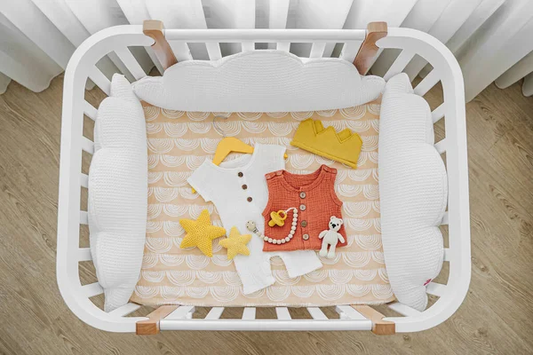 White wooden baby crib with pillows shaped clouds  in baby's room. Newborn clothes and accessories in cot. Top view of child's bed