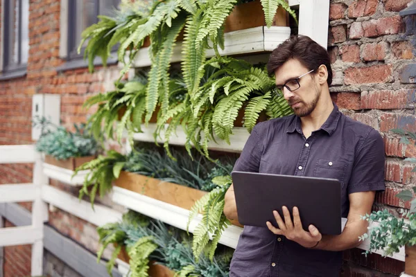 Young entrepreneur working outside using modern laptop. Standing near brick wall, plants, eco office. Successful business people.