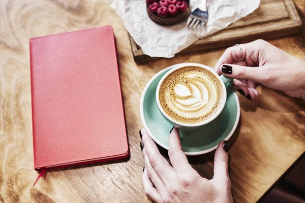 Cup of coffee latte and red book on wooden table or background in woman hands from above.