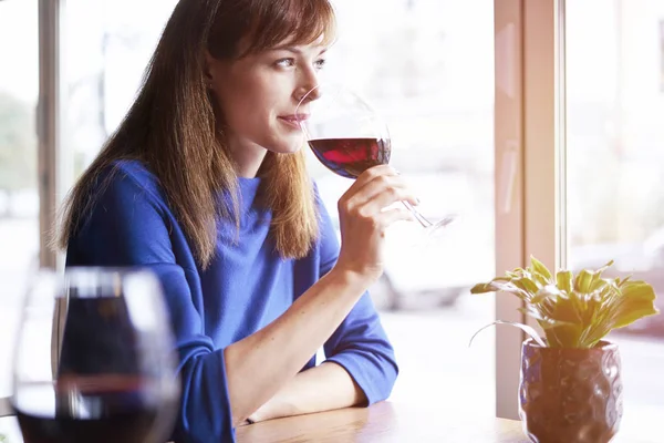 Beautiful woman drinking red wine with friends in cafe, portrait with wine glass near window. Vocation holidays evening bar concept.