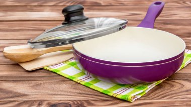 Cam kapaklı mor tava, ahşap mutfak aletleri ve ahşap masada havlu. Yemek pişirme.