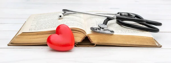 Opened book with stethoscope and red heart on white wooden table.