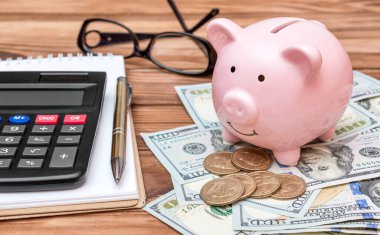 Üzerinde not defteri, hesap makinesi, dolar banknotları ve madeni paraları olan bir kumbara.. 