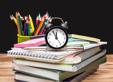 Tahta arka plandaki ahşap masada öğrenci malzemeleri olan saat. Okula gitme zamanı..